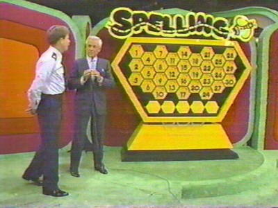 Two men standing in front of a colorful game show set featuring a large hexagonal game board labeled 'Spelling Bee.'
