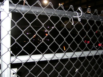 Night view of a chain-link fence with blurred reflections of lights from vehicles or structures behind it.