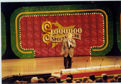 A host stands on stage in a sparkling jacket, addressing an audience in front of a colorful backdrop featuring dollar signs and the words '$1,000,000 Spectacular' during a special episode of a game show.