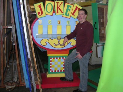A man stands next to a colorful game board titled 'JOKER' featuring slots for numbers and decorative elements, set in a storage area.