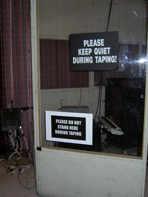 A glass booth with two signs: one instructing to 'Please Keep Quiet During Taping' and the other saying 'Please Do Not Stand Here During Taping'.