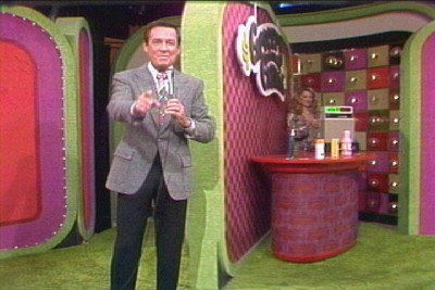 The Grocery Game set, with a contestant in the background, presenting a game on The Price is Right. We see the turntable spin as the host throws to break.