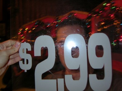 Adam holding a sign displaying the price $2.99, with festive lights blurred in the background.