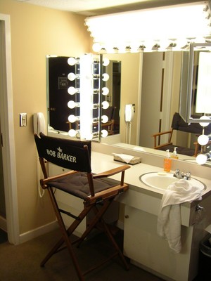 A vanity area in a dressing room featuring a mirror surrounded by bright lights, a wooden chair with 'Bob Barker' branding, and a sink with toiletries.