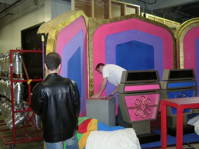 Two individuals working behind the scenes of a game show set, with colorful props in the background.