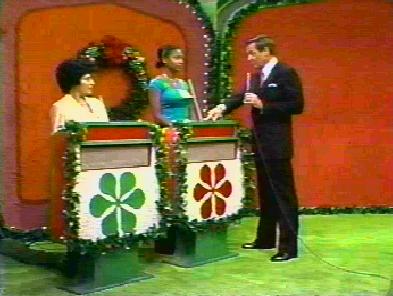 Two contestants stand at showcase podiums decorated with greenery while a host interacts with them in a colorful studio setting.