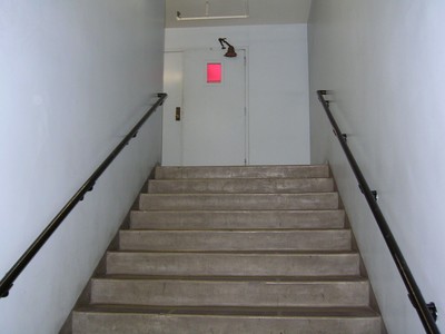A view of a staircase leading up to a door with a red window, featuring black handrails and light-colored walls.