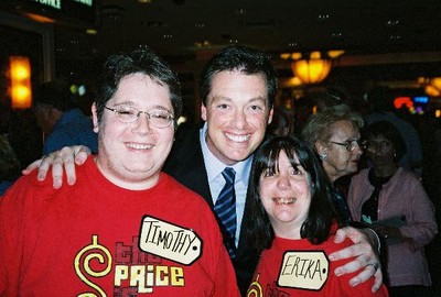 Two fans wearing bright red shirts with name tags posing with a television personality, smiling in a lively setting.