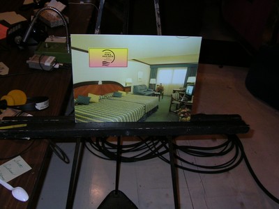 A photo showing a framed image of a hotel room setting on an easel, with a striped bedspread, visible furniture, and a bright light from a window.