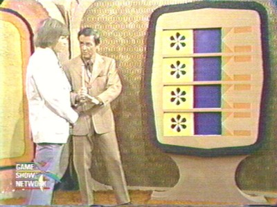 Bob Barker and contestant standing near the Bonus Game, featuring a colorful board with symbols. One man is dressed in a white jacket, while the other is in a brown suit.