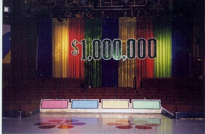 Stage of a game show featuring a '$1,000,000' sign with colorful backdrop and empty podiums.