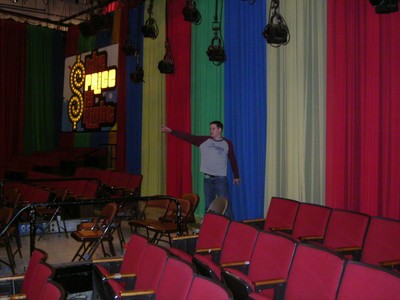 A man stands in a colorful studio set with rows of red chairs, gesturing towards the stage backdrop with the words 'The Price is Right' illuminated.