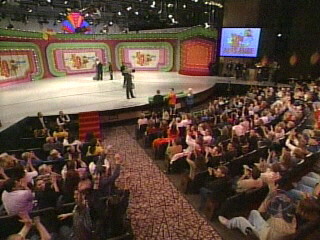 Audience members clapping and cheering during a live taping of a game show on stage, with colorful set design in the background.
