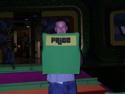 A person smiling while holding a green board with the word 'PRICE' displayed on it, set against a colorful background resembling a game show stage.