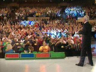 A lively audience cheering and clapping while watching a game show, with a host speaking on stage.