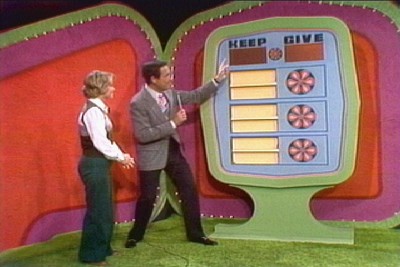 A game show host points at a large game board labeled 'KEEP' and 'GIVE' while a contestant stands beside him on a colorful set.