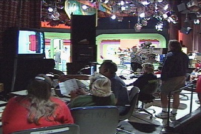 A behind-the-scenes view of a television studio with crew members at work, monitors displaying the show, and colorful set pieces in the background.