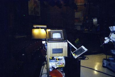 A vintage control station with a monitor and various buttons, surrounded by studio equipment in a dimly lit setting.