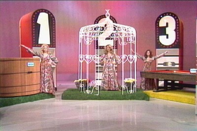 Three female hosts in vintage outfits stand next to a Most Expensive setup on a brightly colored stage, with large numbers indicating prize options.