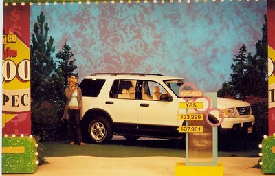 A contestant stands next to a white SUV on a game show set, with a colorful backdrop and a game display showing different prize amounts.