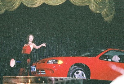 A woman in a red dress poses beside a red car on stage, with a black curtain and decorative gold drapes in the background.