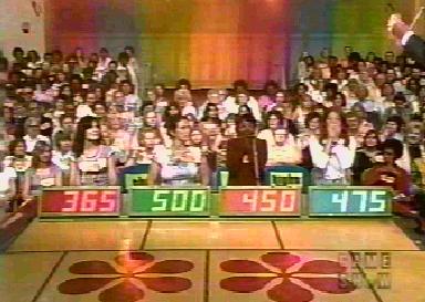 A crowded audience at a game show, with contestants displaying their scores on brightly colored screens. The background features a colorful set design.