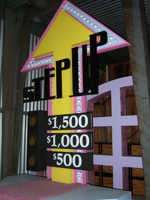 A bright yellow and pink game show set piece with an upward arrow and the word 'STEP UP' prominently displayed. Below, there are three price points: $1,500, $1,000, and $500.