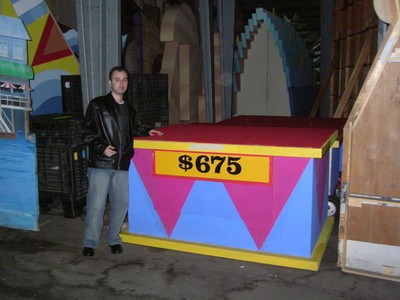 A person standing next to a colorful Easy as 123 game board marked with the price $675, set in a storage area filled with props.