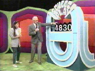 The Squeeze Play board displaying the number 4830, while a contestant stands beside him, both in front of a colorful game show backdrop.