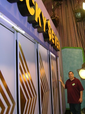 A man standing next to a large sign that reads 'LUCKY SEVEN' in a brightly lit studio setting, showcasing a game show backdrop.