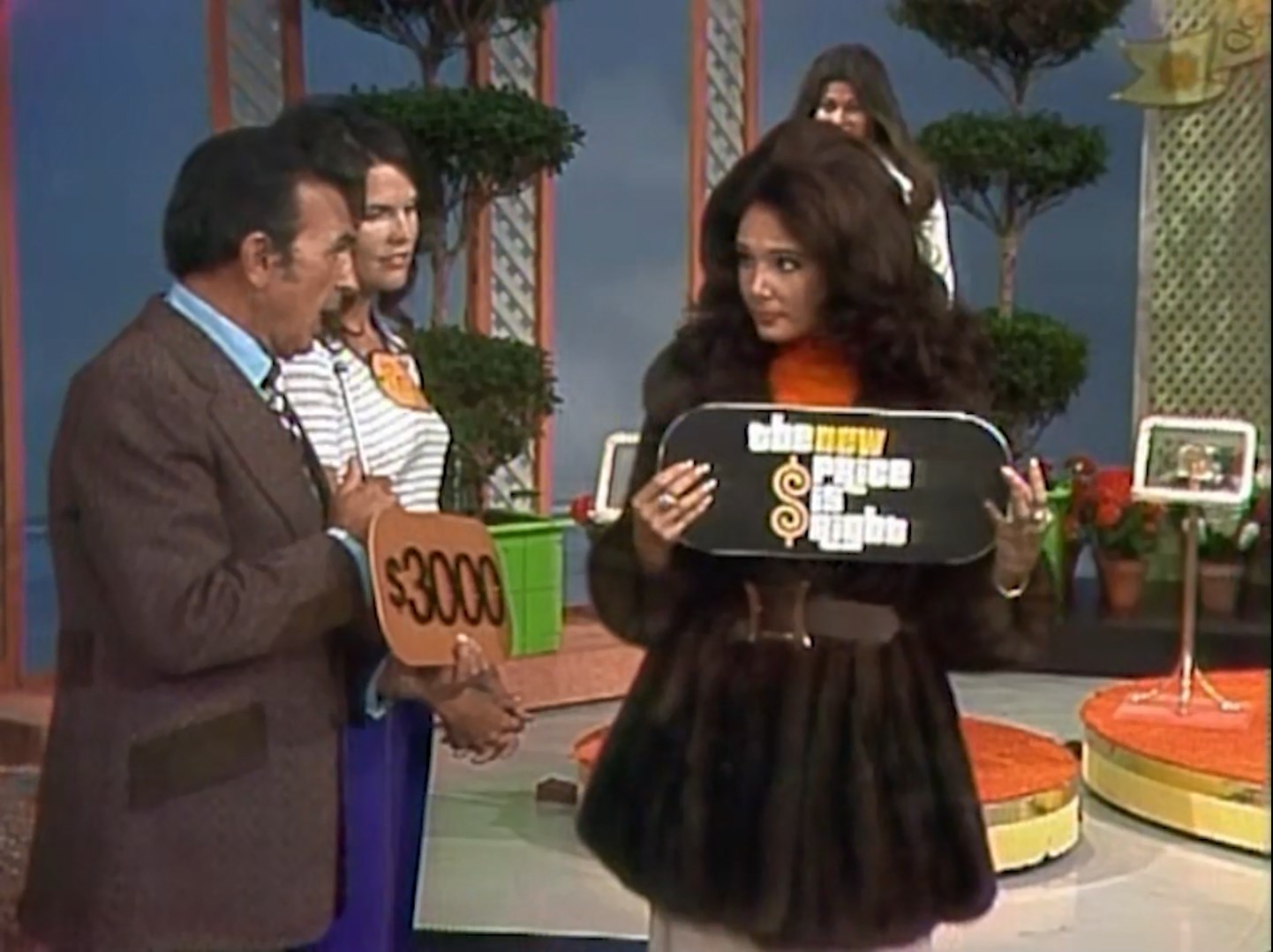 A game show contestant holds a price tag reading 00 while a female model presents a sign that says 'the new price is right'. The background features potted plants and colorful game show set decor.