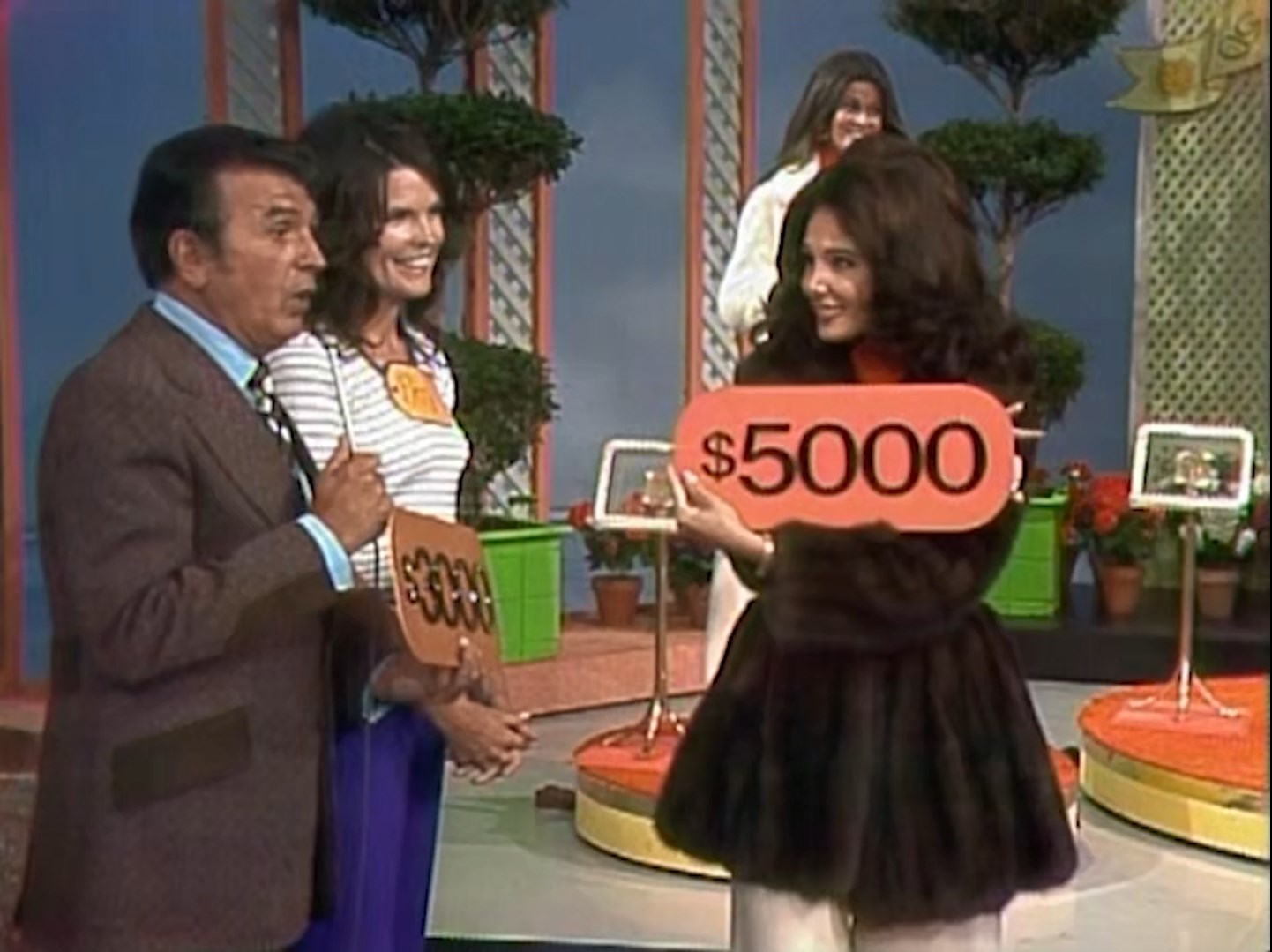 A game show scene featuring a host and two contestants. One contestant is holding a sign indicating a prize of 00 while another contestant stands beside her with a lower value sign. The setting includes potted plants and game show decorations.