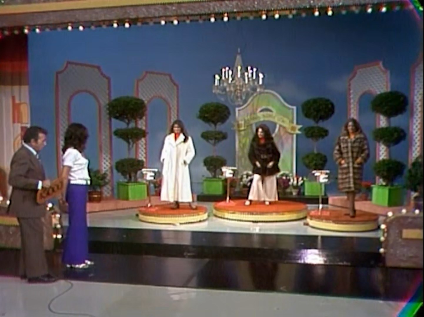 A game show set featuring three models on circular podiums showcasing various coats, with a host and contestant watching intently.