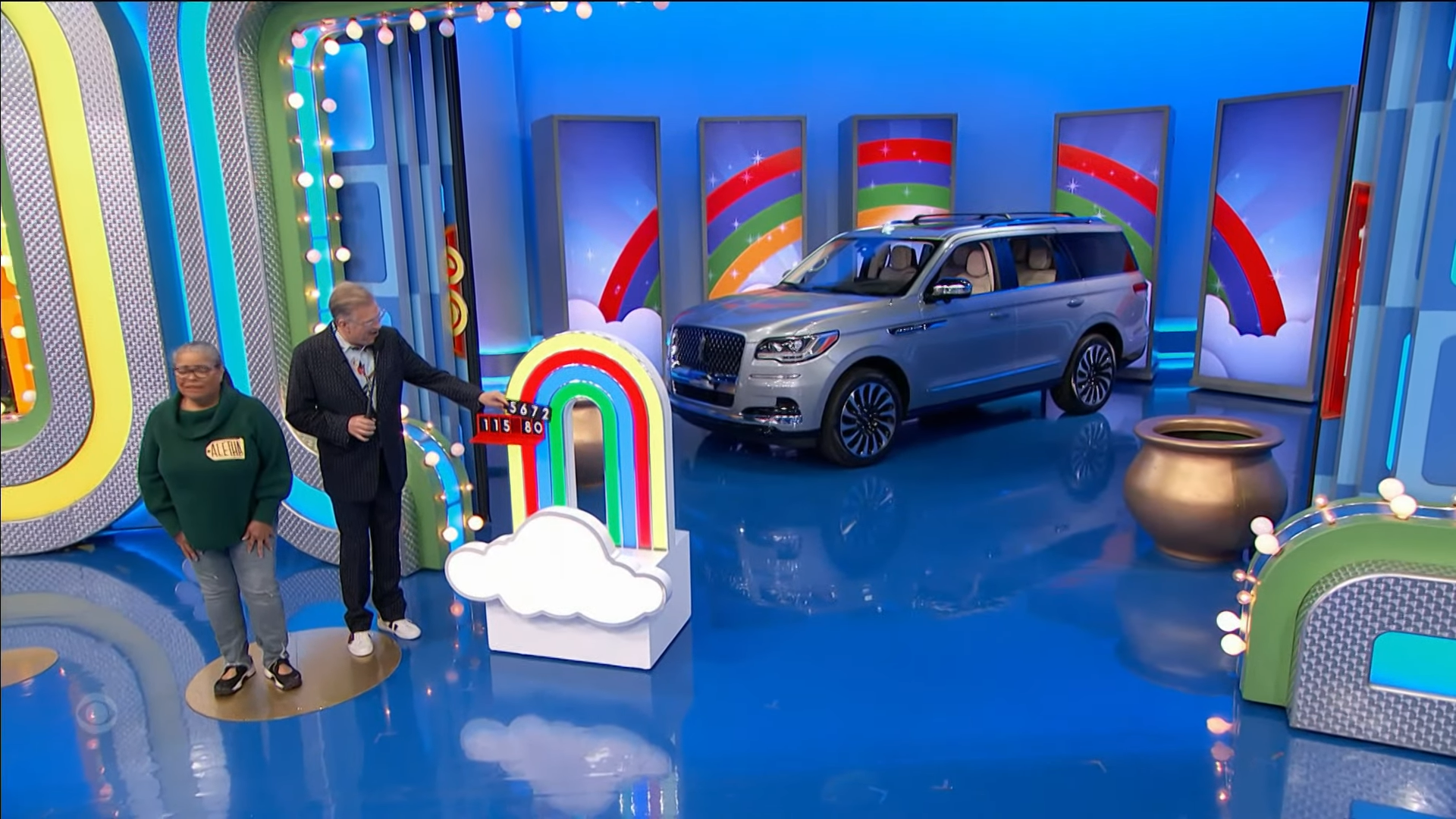 Contestant Aletha stands on the stage of the game show, next to a colorful prize display and a silver luxury SUV.