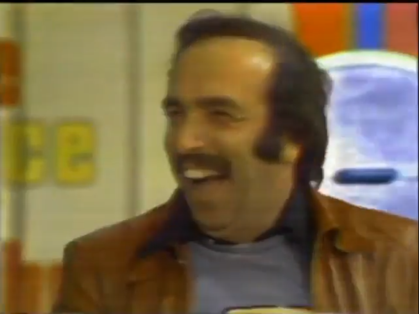 A contestant smiling excitedly while participating in a game show, wearing a brown leather jacket and a blue shirt, with a colorful backdrop behind him.