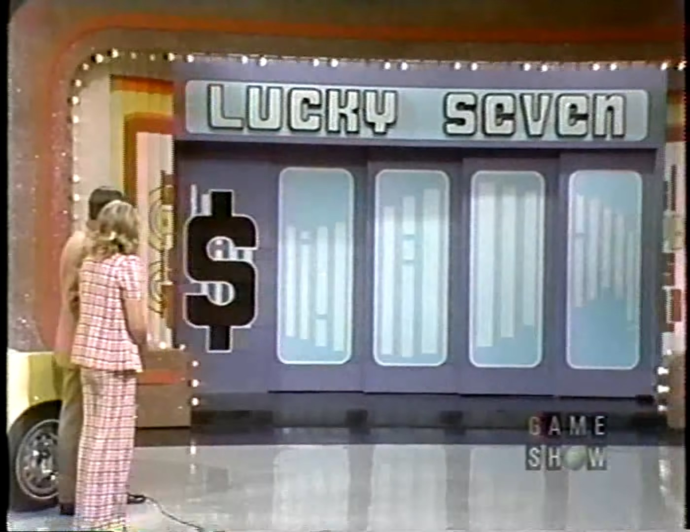 Contestants prepare to play the Lucky Seven game on a game show stage featuring a large dollar sign graphic.