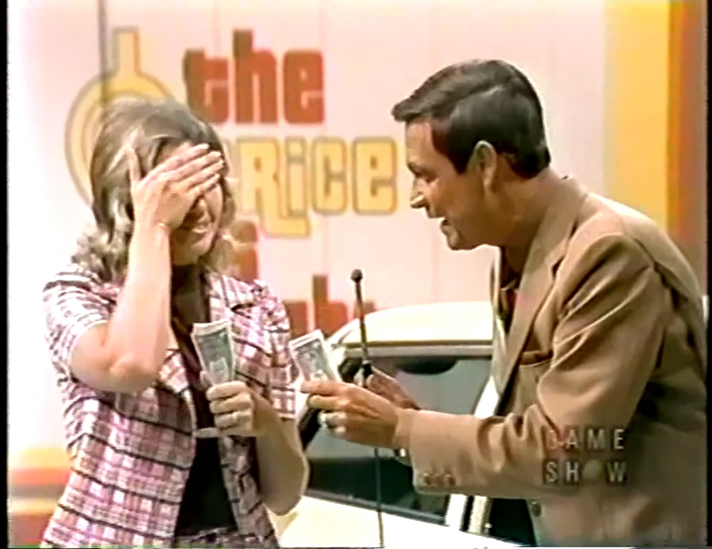 A contestant on a game show holds cash while interacting with the host, both are engaged and expressive against a colorful game show backdrop.