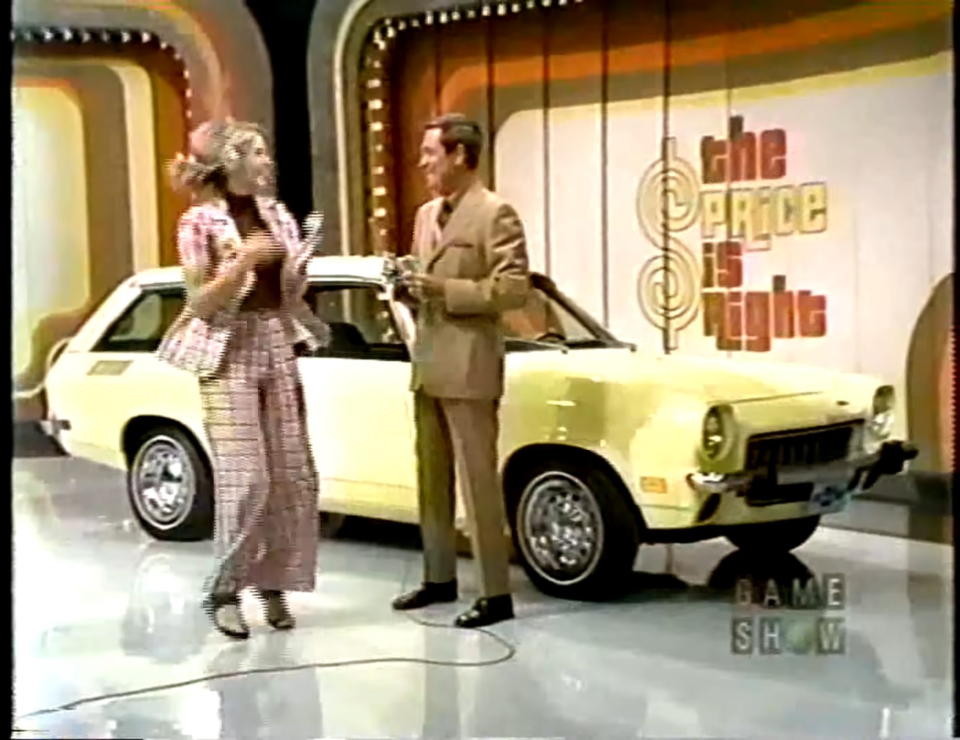 A female contestant and a male host are celebrating next to a yellow car on the set of 'The Price is Right' in 1973. The background features colorful stage decorations.