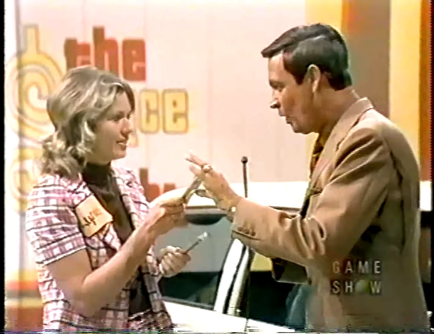 A female contestant smiles while holding a pen and interacting with the host on stage during a game show.