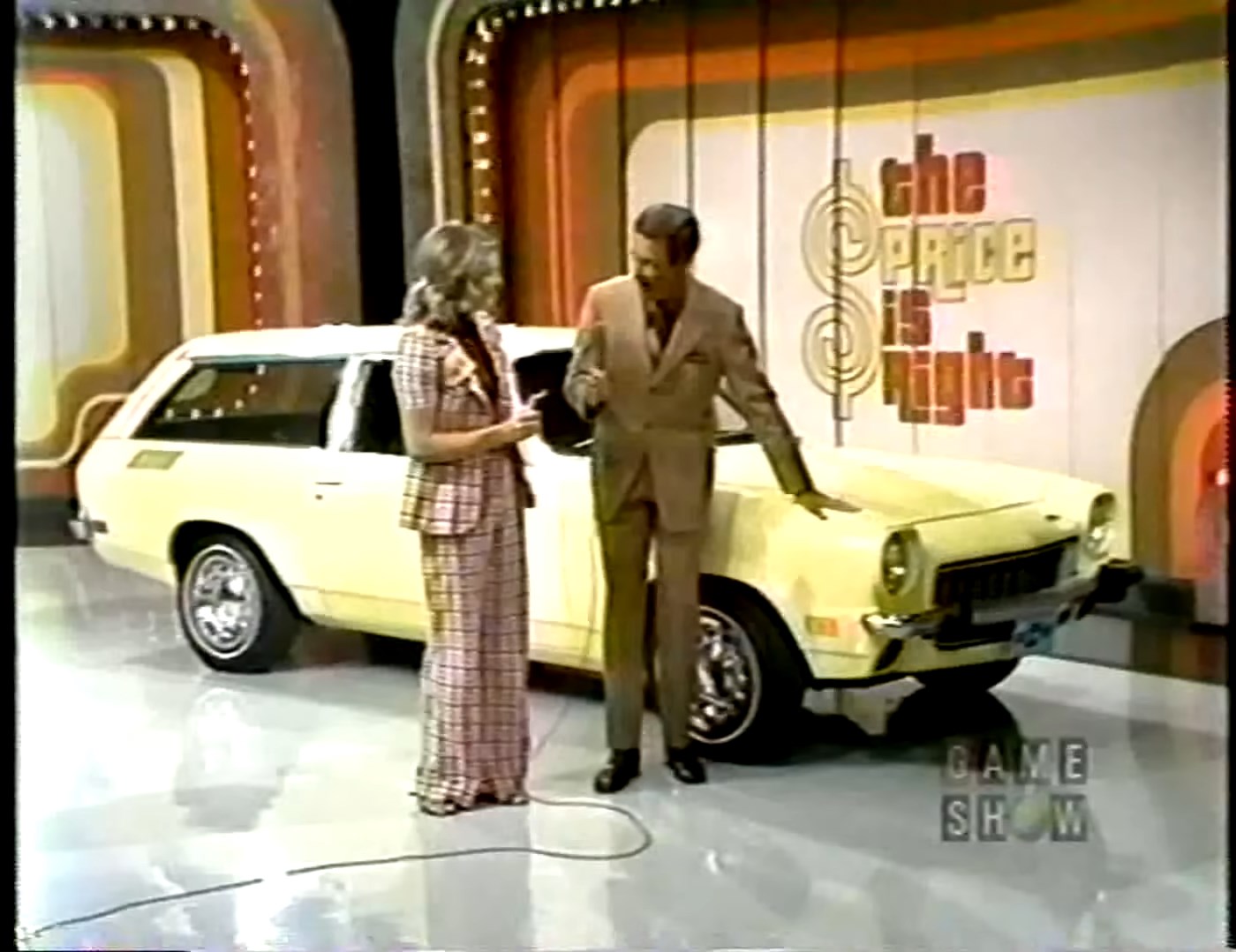 A game show host stands next to a contestant and a yellow car during a segment of 'The Price is Right'. The backdrop features colorful game show graphics.