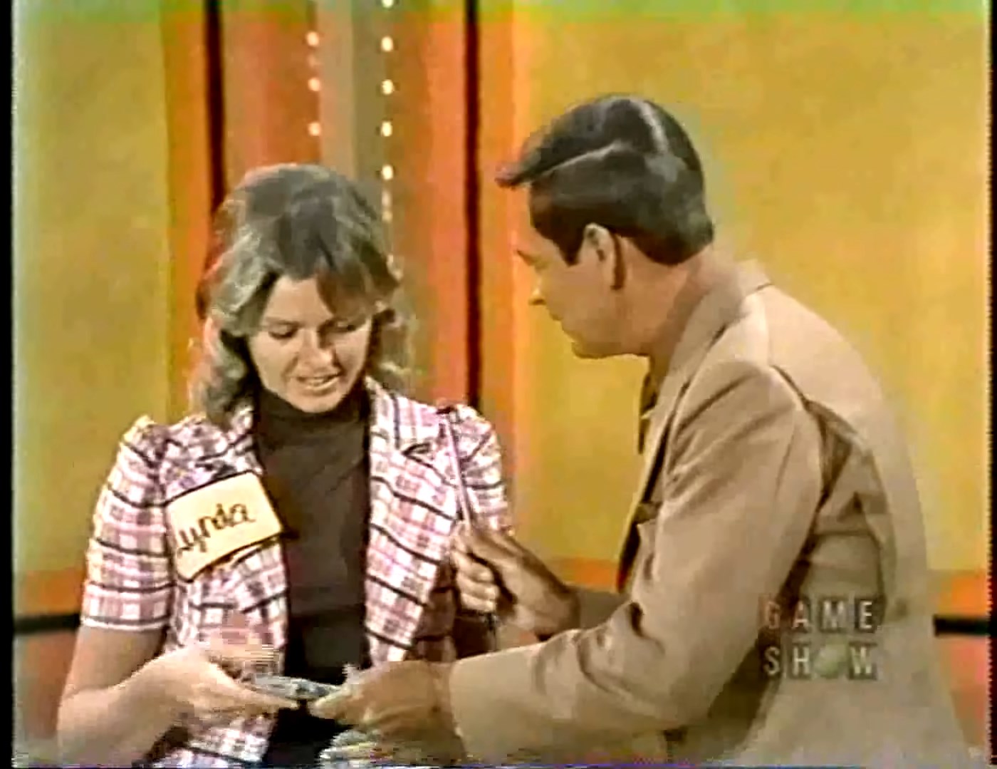 A game show contestant named Lynda looks disappointed as she holds cash, preparing to play the game Lucky Seven.