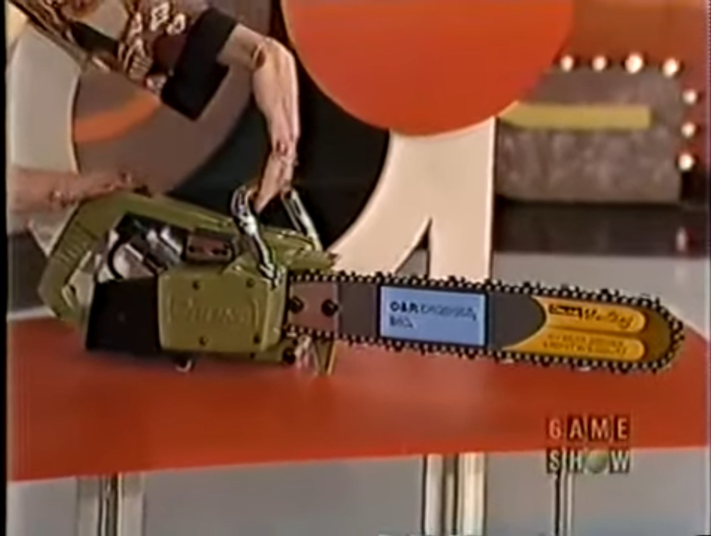A woman holds a green chainsaw on a game show set, showcasing it as a potential prize.