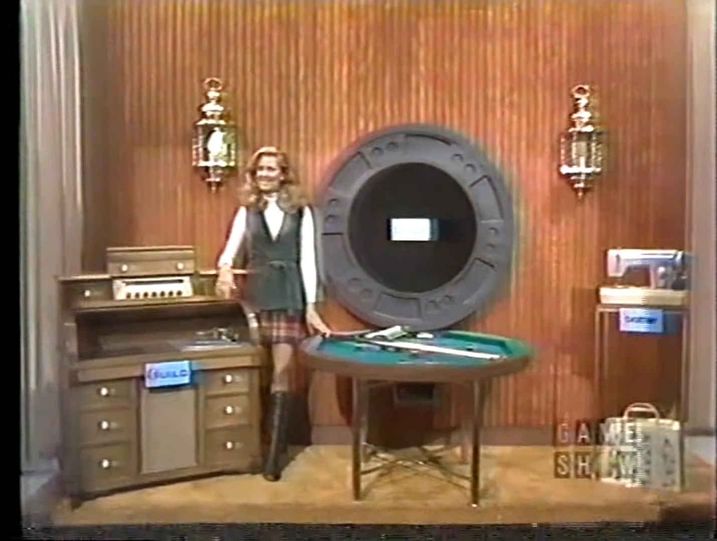 A game show set featuring a presenter beside a wooden cabinet, a roulette-style game wheel, and a gambling table, with decorative lanterns in the background.