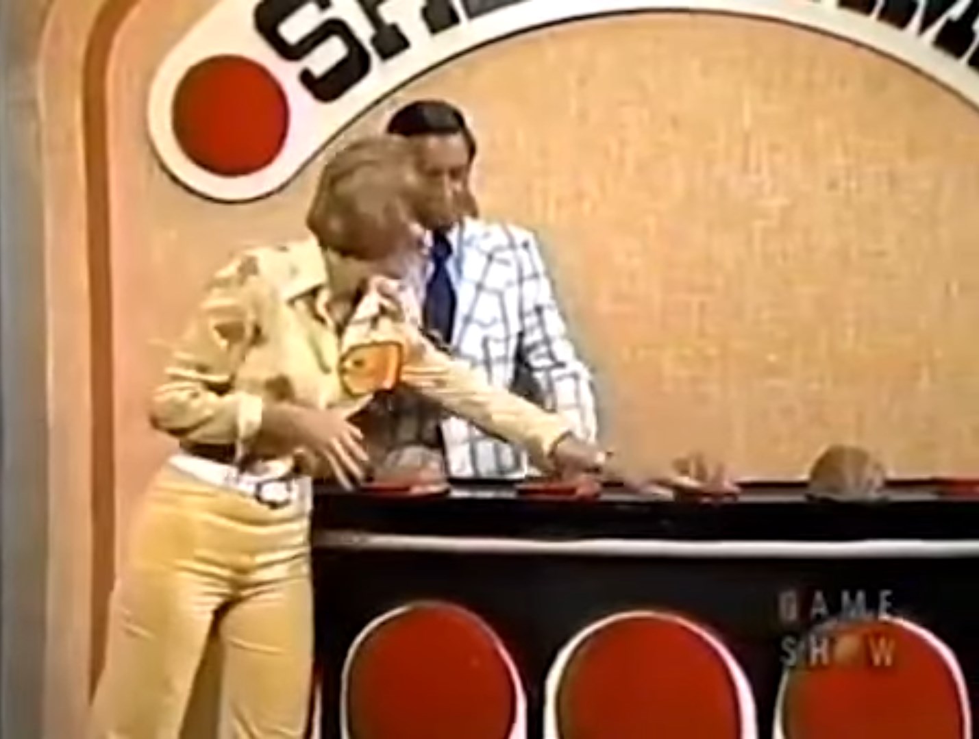 A contestant selects a shell in the Shell Game while a host observes, with a colorful game show backdrop.