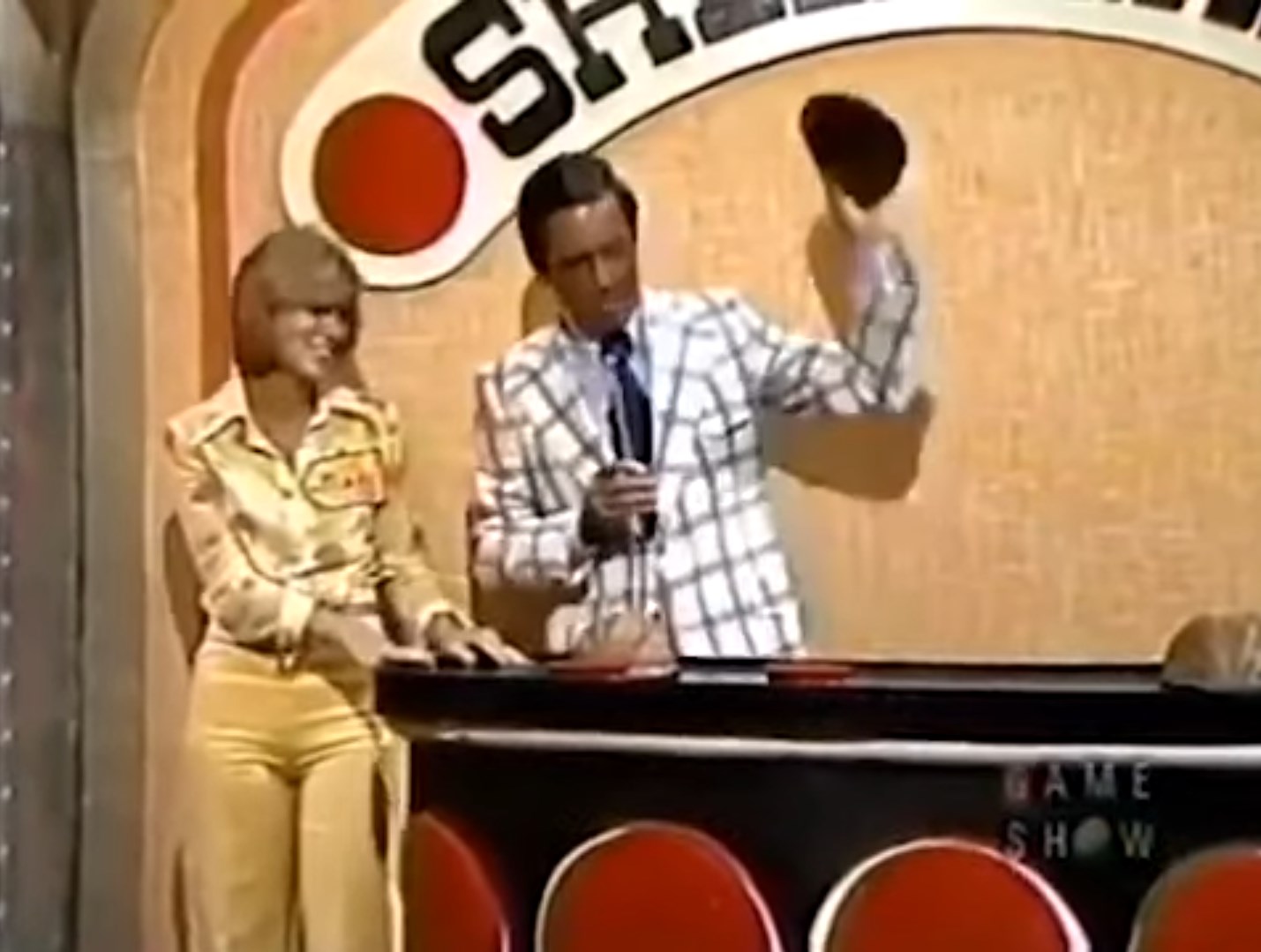 A game show host presenting the Shell Game on stage, wearing a plaid suit and holding a shell, while a contestant smiles beside him.