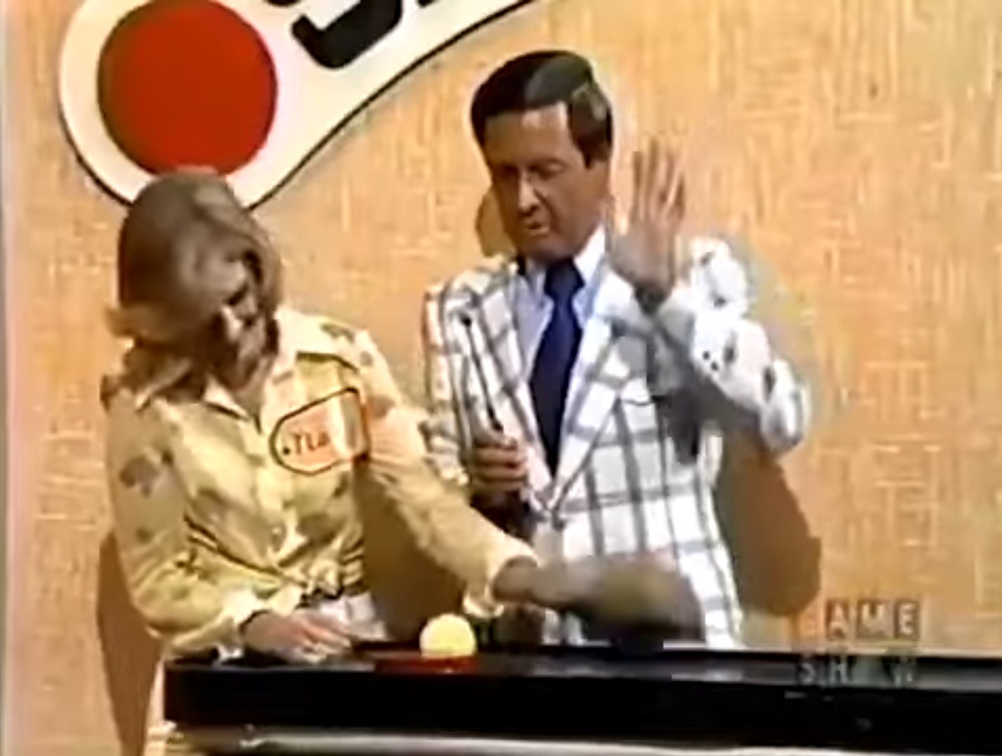 A woman in a yellow shirt participates in the Shell Game on stage, placing a shell as a game host gestures while explaining rules.