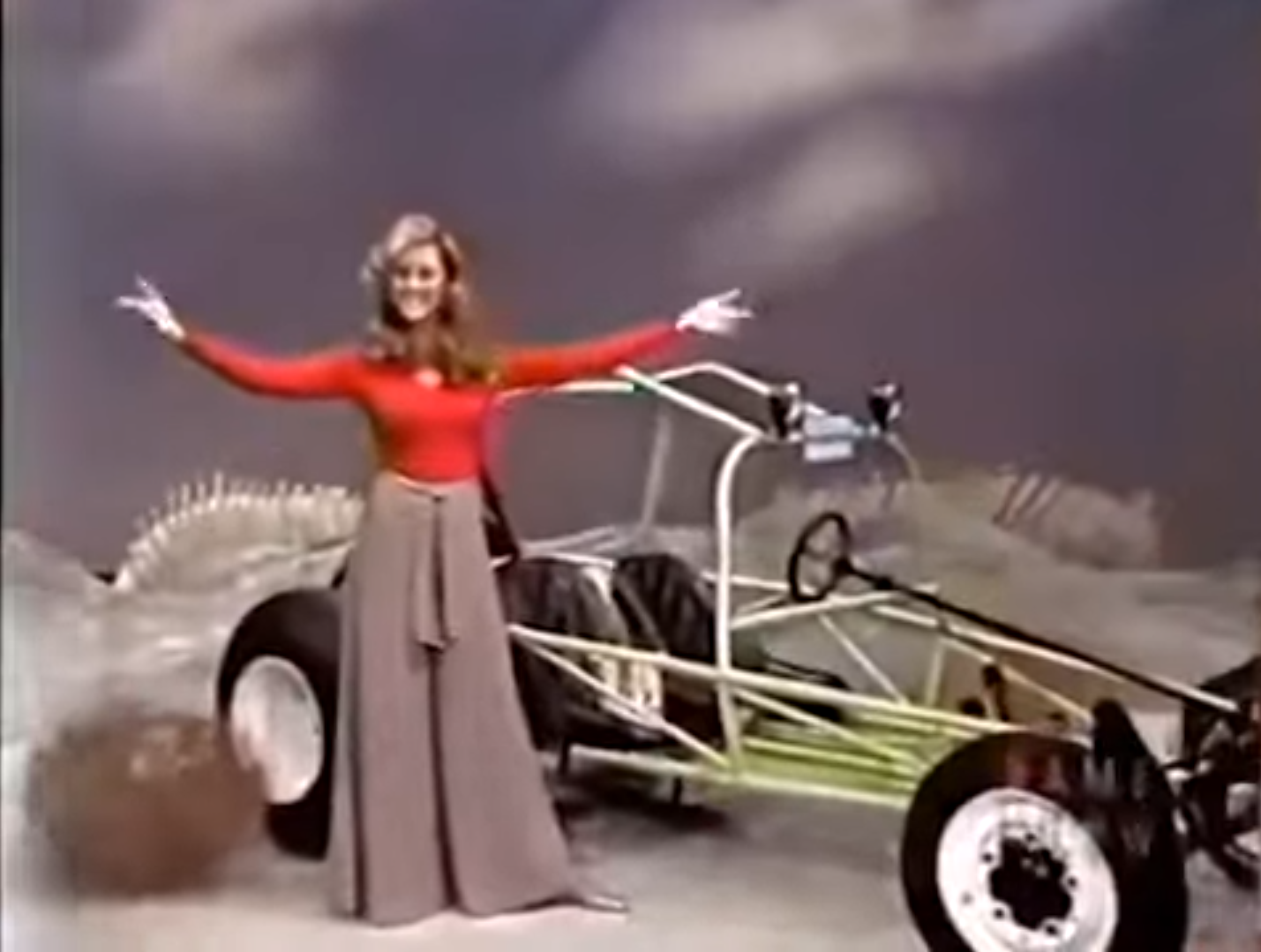 A woman in a red top and long skirt joyfully presents a dune buggy on stage.