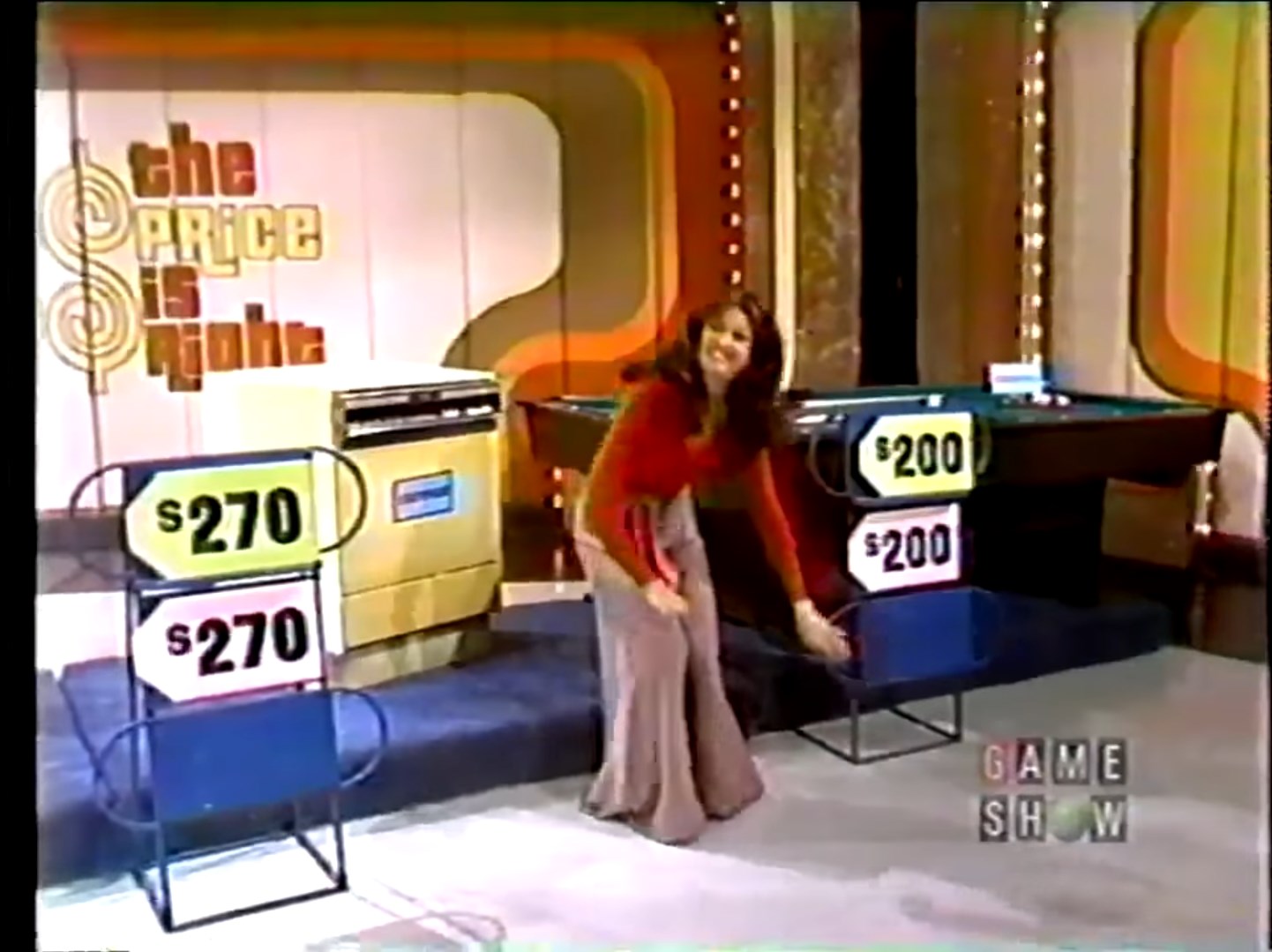 A contestant in a red top and long skirt arranges price tags on stands while smiling, with prizes and a game show set in the background.