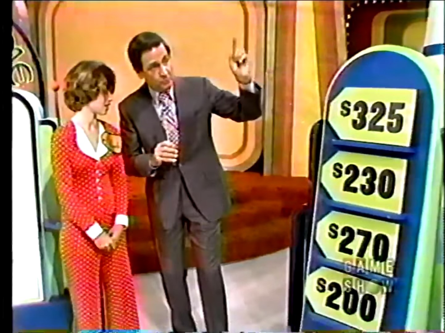 A game show host explains the rules of the Race Game to a contestant, with price tags of 5, 0, 0, and 0 displayed on a board.
