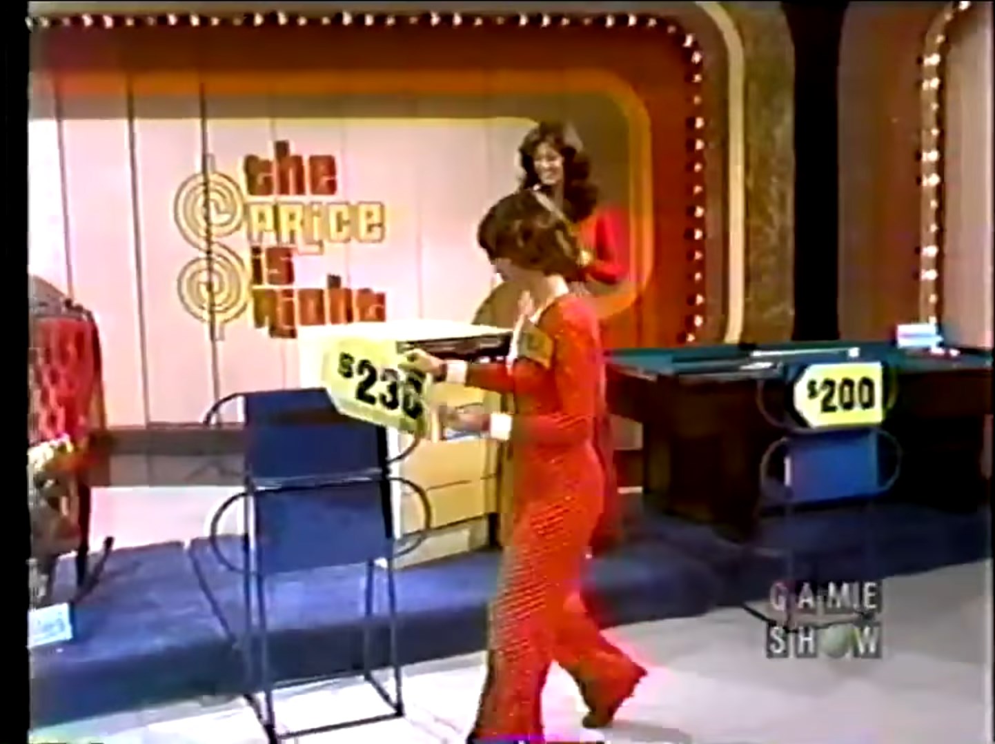 A contestant dressed in a red outfit is holding a price tag marked 0 while placing it on a prize in 'The Price is Right' game show. A model claps in the background.